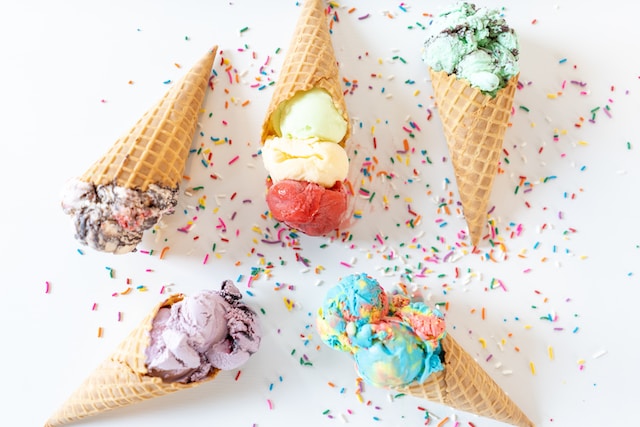 5 ice cream or gelatos in wafer cones. Each different colours and flavours. Laying on a white back ground and surrounded by multicoloured sprinkles.